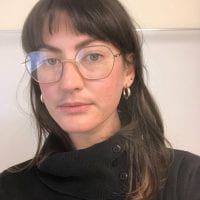 light skinned woman with long dark hair and bangs wearing large metal-rimmed glasses