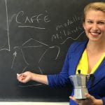 woman with light skin and blond hair pulled into a ponytail holding an old espresso pot in front of a chalkboard