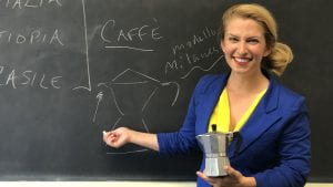 woman with light skin and blond hair pulled into a ponytail holding an old espresso pot in front of a chalkboard
