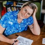 A white woman with short blond hair with dark roots, wearing glasses and smiling. Drawing a cartoon.