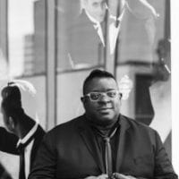 Black and white photo fo a black man wearing glasses and a suit jacket.