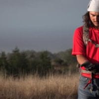 A person standing in a field wearing a red tee shirt and bandana on head. They have long brown hair. They are looking down at soil in their hands.