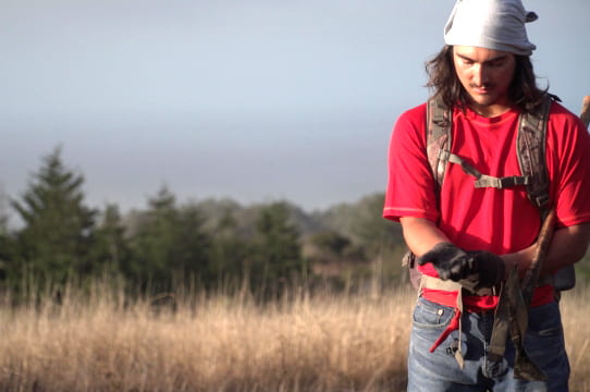 a young man with long hair and a bandana on his head wearing a red tee shirt is holding soil in his hands and looking at it. He is standing in a field of tall grass.