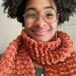 woman with medium brown skin dark curly hair in an afro wearing glasses and a thick orange scarf