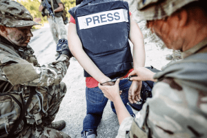 soldiers wearing camouflage detaining a person wearing a vest with "press" printed on it